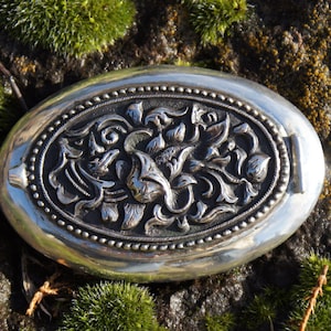 May include: Oval silver compact with ornate floral relief. The compact features a detailed, raised floral design within an oval frame, surrounded by a beaded border. The compact is set against a backdrop of green moss and a dark, textured surface.