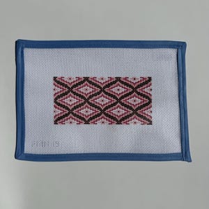 May include: A rectangular needlepoint canvas with a geometric pattern in shades of pink, black, and white. The design is framed by a blue border. The canvas is likely used for creating decorative items or artwork.