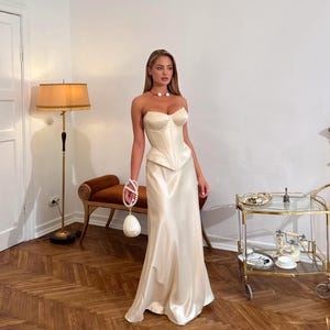 May include: A cream-colored strapless corset top paired with a matching floor-length satin skirt. The outfit is accessorized with a pearl choker necklace and a small, round, white handbag featuring a pearl-beaded handle.