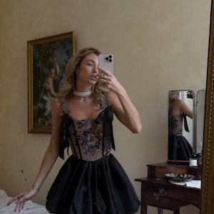 May include: A sleeveless corset top featuring a dark floral pattern with black ribbon shoulder straps and a matching black bubble skirt. The corset has a structured bodice with vertical boning, and the skirt has a voluminous, rounded silhouette.