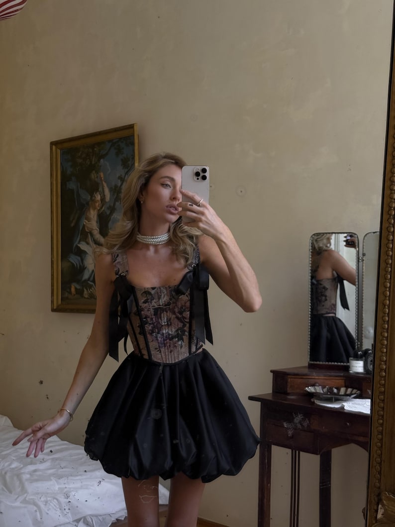 May include: A sleeveless corset top featuring a dark floral pattern with black ribbon shoulder straps and a matching black bubble skirt. The corset has a structured bodice with vertical boning, and the skirt has a voluminous, rounded silhouette.