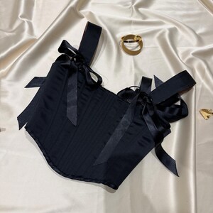 May include: A black corset top featuring structured vertical seams and wide shoulder straps adorned with decorative satin ribbon bows at the front. The garment is crafted from a smooth, solid black fabric with a subtle sheen throughout.