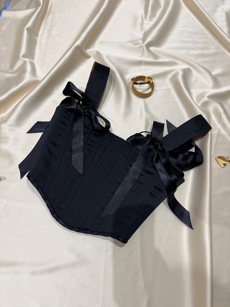 May include: A black corset top featuring structured vertical seams and wide shoulder straps adorned with decorative satin ribbon bows at the front. The garment is crafted from a smooth, solid black fabric with a subtle sheen throughout.