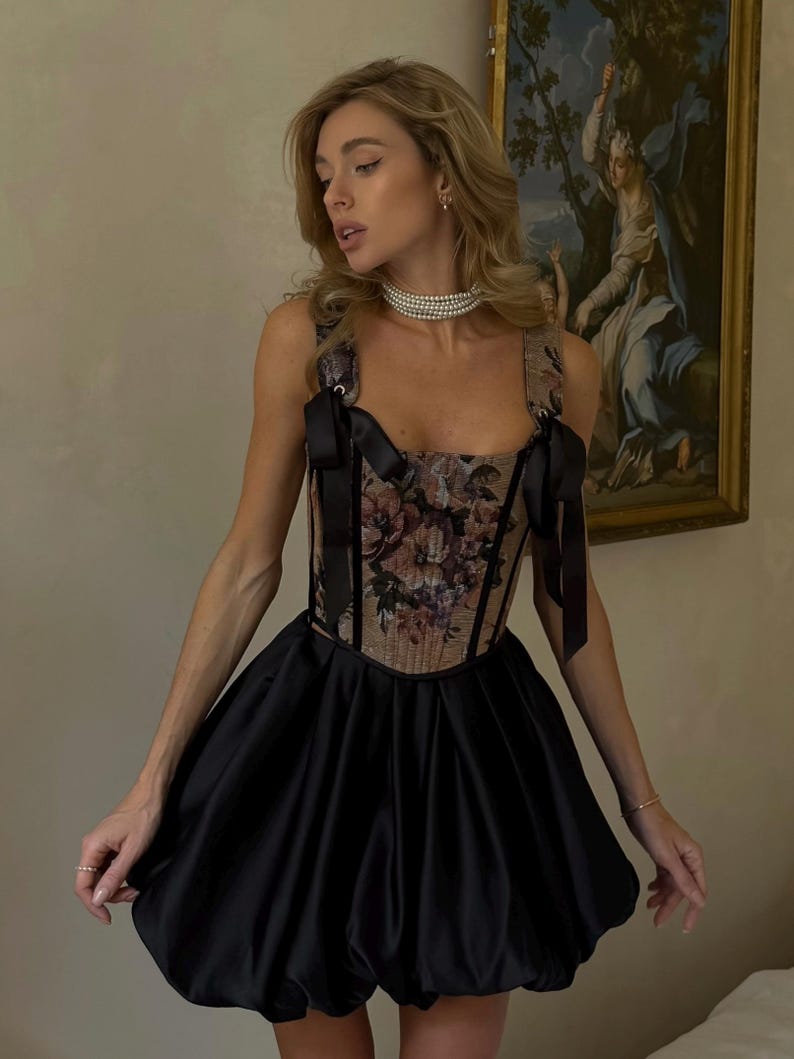 May include: A sleeveless dress featuring a structured corset-style bodice with a floral tapestry pattern, black ribbon shoulder ties, and a voluminous black satin bubble skirt that falls above the knee, creating a distinct silhouette.
