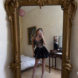 May include: A black mini dress featuring a structured corset-style bodice with intricate floral embroidery and thin shoulder straps with bows. The skirt is voluminous and puffed, ending above the knees, paired with black pointed-toe heels.