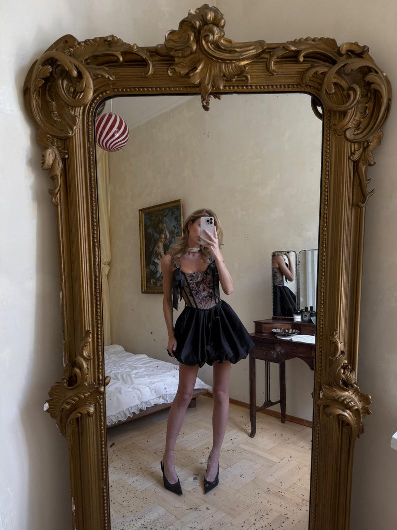 May include: A black mini dress featuring a structured corset-style bodice with intricate floral embroidery and thin shoulder straps with bows. The skirt is voluminous and puffed, ending above the knees, paired with black pointed-toe heels.