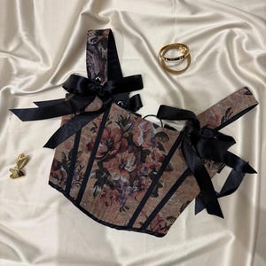 May include: A corset-style top featuring a floral tapestry pattern in muted tones of brown, red, and green. The garment is accented with black vertical piping and black satin ribbon ties at the shoulders. Two gold-toned metal rings and a small gold heart-shaped charm are placed nearby.