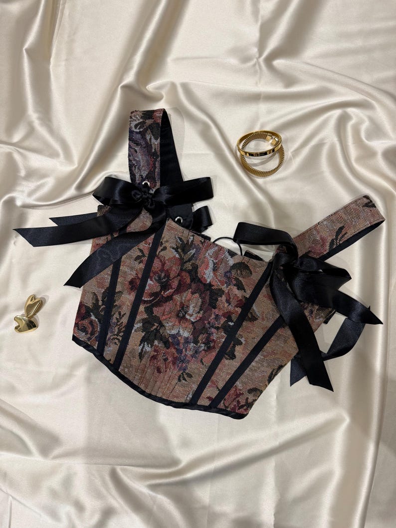 May include: A corset-style top featuring a floral tapestry pattern in muted tones of brown, red, and green. The garment is accented with black vertical piping and black satin ribbon ties at the shoulders. Two gold-toned metal rings and a small gold heart-shaped charm are placed nearby.