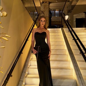 Könnte beinhalten: Ein trägerloses, schwarzes Abendkleid mit herzförmigem Ausschnitt. Das bodenlange Kleid wird mit einer kleinen schwarzen Clutch mit Kettenriemen kombiniert. Die Person steht auf einer Marmortreppe.