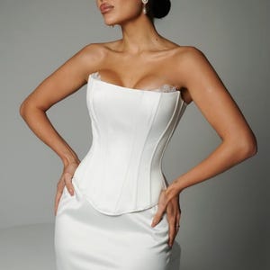 May include: A white satin corset top and matching skirt. The strapless corset has vertical seams and lace trim. The skirt is fitted with a lace hem. The outfit is elegant and suitable for special occasions. The measurements are in centimetres.