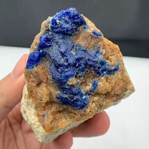 Puede incluir: Un espécimen mineral natural que presenta un llamativo contraste de colores. La pieza muestra una vibrante formación mineral azul profundo sobre un fondo de roca marrón y beige terroso. La textura parece áspera y cristalina.