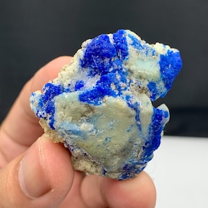 Puede incluir: Un espécimen de lapislázuli azul intenso con motas doradas. La piedra muestra su forma natural con una matriz gris clara. La estructura cristalina del mineral es visible.