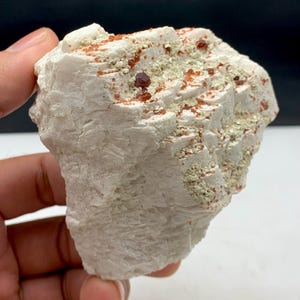 Puede incluir: Un espécimen mineral blanco rugoso con pequeños cristales de color marrón rojizo y parches de un material más claro, de color beige. La roca tiene una superficie texturizada y se sostiene en una mano, mostrando su apariencia natural y en bruto.