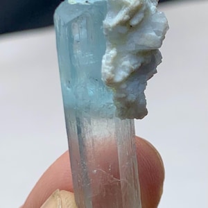 Puede incluir: Un cristal de aguamarina translúcido de color azul claro con una formación mineral blanca y texturizada adherida. El cristal se sostiene en una mano, mostrando su forma geométrica natural y variaciones de color. La formación mineral es de color beige claro.