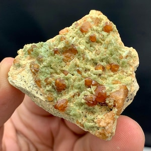 Puede incluir: Primer plano de un espécimen mineral con una matriz de cristales verde claro y grupos de cristales de color marrón anaranjado. El mineral tiene una superficie rugosa y texturizada. El fondo es oscuro.