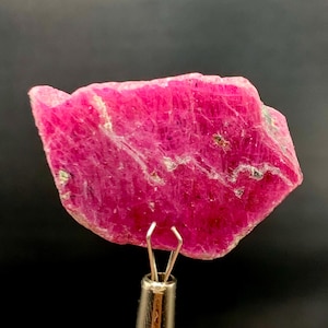 May include: A vibrant, raw ruby gemstone, showcasing a deep pink hue with natural textures and subtle white veining. The gemstone is held by a silver metal tool against a dark background. This mineral specimen is ideal for collectors.