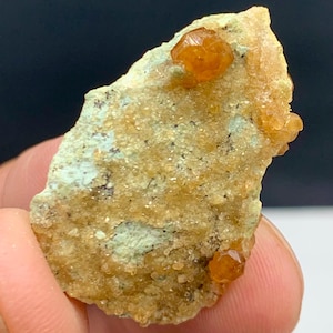 Puede incluir: Un espécimen mineral en bruto con una matriz verde claro y beige. Varios pequeños cristales de color marrón anaranjado están incrustados en la roca. La superficie del espécimen es rugosa y texturizada. Fondo oscuro.