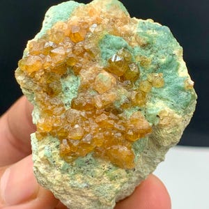 Puede incluir: Un espécimen mineral natural con grupos de cristales de color naranja dorado sobre una matriz de roca verde y gris. Los cristales tienen una estructura cristalina brillante. La apariencia general es áspera y texturizada, mostrando la belleza en bruto de las formaciones terrestres.
