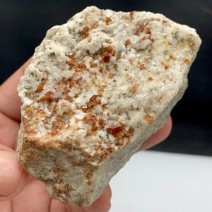 Puede incluir: Un espécimen mineral natural y rugoso con una matriz de piedra gris claro y blanca. La superficie está salpicada de pequeños cristales de color marrón rojizo, creando una apariencia texturizada. El mineral se sostiene en una mano.