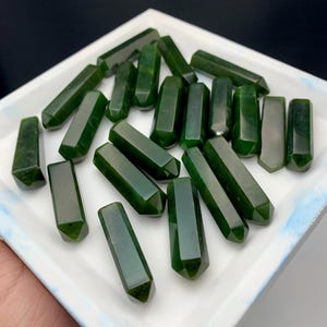 May include: A collection of dark green, hexagonal-shaped gemstone wands. The wands are arranged on a white tray, showcasing their polished surfaces and varying sizes. The wands have a deep, rich green color, with some showing slight variations in tone.