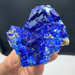 Puede incluir: Un vibrante espécimen mineral azul con una estructura cristalina. La piedra presenta un color azul profundo y saturado con motas doradas y blancas. La superficie rugosa y texturizada del mineral es visible, mostrando su forma natural. El fondo es gris oscuro.