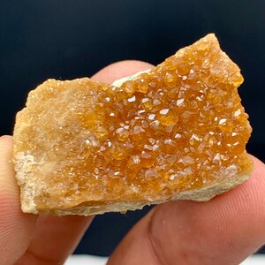 Puede incluir: Primer plano de un grupo de pequeños cristales de color amarillo anaranjado brillante. Los cristales están densamente empaquetados sobre una base rugosa de color beige. La formación mineral es sostenida por los dedos de una persona sobre un fondo oscuro.