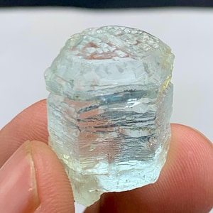 May include: A translucent, light blue aquamarine crystal with a rough, textured surface. The gemstone is held between fingers, showcasing its natural, unpolished form. The crystal's facets catch the light, revealing subtle color variations.