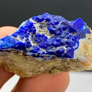 Puede incluir: Una gema de lapislázuli en bruto y natural. La piedra presenta un color azul profundo con estructuras cristalinas y motas de pirita dorada, sobre una matriz beige. La forma rugosa e irregular resalta su forma natural.