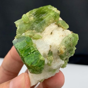 Puede incluir: Un espécimen mineral en bruto y natural con cristales verdes vibrantes incrustados en una matriz blanca. Los cristales tienen un brillo vítreo y están agrupados, creando un contraste llamativo con el fondo blanco. El mineral se sostiene en una mano.