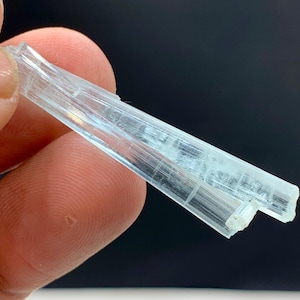 May include: A close-up of a pale blue crystal formation. The crystal is held between fingers and appears translucent with visible internal structures. The background is a dark, solid color, highlighting the crystal's details. The crystal is a natural mineral specimen.