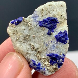 Puede incluir: Un trozo de piedra de lapislázuli en bruto con una matriz gris claro. La piedra presenta parches azul oscuro y motas de pirita dorada. Se aprecia la forma rugosa e irregular de la piedra.