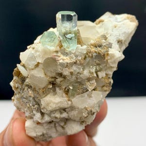 May include: A natural aquamarine crystal cluster. The focal point is a large, clear, light blue-green aquamarine crystal, accompanied by smaller crystals. The matrix is a mix of white, tan, and brown minerals, creating a textured appearance.