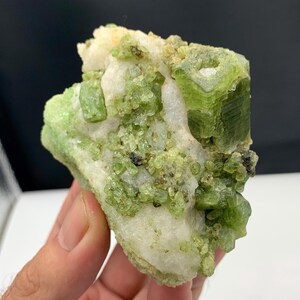 Puede incluir: Primer plano de un espécimen mineral natural en bruto. La piedra presenta una mezcla de colores verde claro, verde lima y blanco. El mineral tiene una estructura cristalina con facetas visibles y una textura rugosa. También hay pequeñas inclusiones negras.