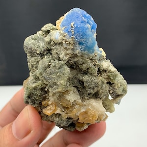 Puede incluir: Primer plano de un espécimen mineral con una llamativa formación de cristal azul. La matriz está compuesta por minerales verdes, grises y dorados. La muestra se presenta sobre un fondo neutro, resaltando sus colores naturales.