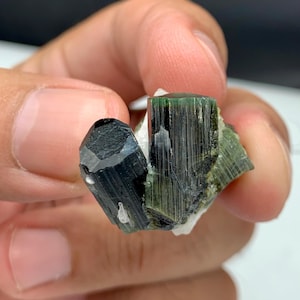 May include: A cluster of dark green and black tourmaline crystals. The crystals have a prismatic structure with a glassy lustre. The tourmaline is set against a white matrix. The overall appearance is of a natural, raw gemstone.