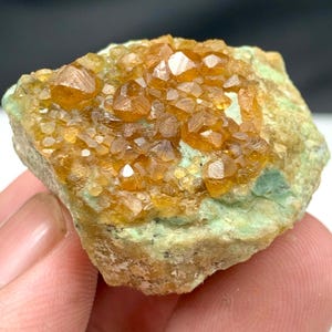 Puede incluir: Espécimen mineral natural con grupos de cristales de color naranja dorado sobre una matriz verde claro. Los cristales tienen una apariencia brillante, similar a una gema, que contrasta con la base terrosa. La textura general es áspera y desigual.