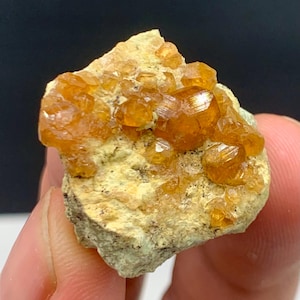 Puede incluir: Primer plano de un espécimen mineral natural. El espécimen presenta grupos de pequeños cristales de color marrón anaranjado sobre una matriz de color claro. Los cristales tienen una calidad brillante y reflectante, y la matriz tiene un aspecto rugoso y texturizado.