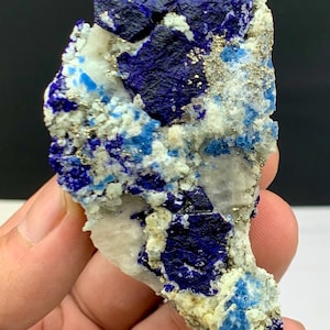 Puede incluir: Primer plano de un espécimen de lapislázuli. La piedra presenta colores azul oscuro, azul claro y blanco con motas doradas. El mineral se sostiene en una mano sobre un fondo negro.