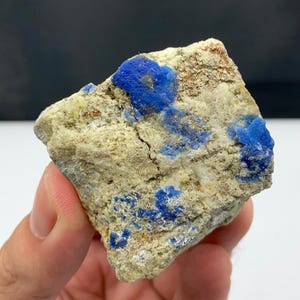 Puede incluir: Una roca rugosa de forma irregular, con una base de color beige claro. Presenta parches de depósitos minerales de color azul vibrante. La textura natural y las variaciones de color de la roca son visibles. La roca se sostiene en una mano.