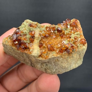 Puede incluir: Una formación rocosa natural con una base verde claro, cubierta de grupos de pequeños cristales de color marrón rojizo y brillantes. Los cristales tienen una apariencia facetada, captando la luz. La textura general es rugosa y natural, mostrando un espécimen mineral.