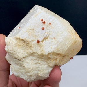 Puede incluir: Un espécimen mineral cristalino de color claro con una gran formación de cristal angular blanquecina. Pequeños cristales de color marrón rojizo están dispersos por la superficie. La base del mineral es de color beige rugoso y texturizado.