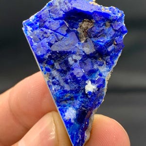 Puede incluir: Un trozo triangular de lapislázuli en bruto, una roca metamórfica azul oscuro. La piedra tiene una superficie rugosa y texturizada con motas blancas y doradas. El mineral se sostiene en una mano sobre un fondo oscuro.