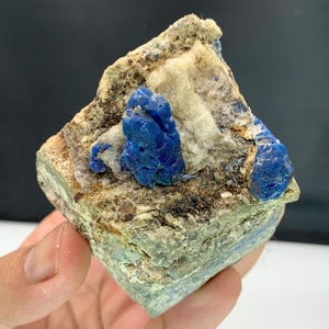 Puede incluir: Un espécimen mineral natural con vibrantes cristales de azurita azul incrustados en una matriz de roca beige y blanca. Los cristales de azurita tienen un color azul intenso, que contrasta con los tonos más claros de la piedra circundante. El espécimen es áspero y texturizado.