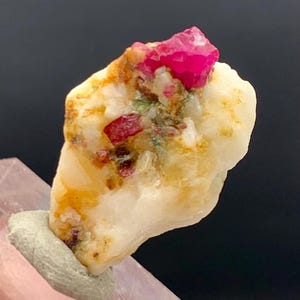 Op de afbeelding: Een natuurlijk mineraal specimen met een lichtgekleurde matrix en heldere rode en roze kristalformaties. Het specimen is gemonteerd op een grijze basis, die de ruwe schoonheid van de edelsteen benadrukt.