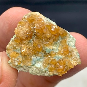 Puede incluir: Primer plano de un espécimen mineral natural en bruto. El espécimen presenta grupos de pequeños cristales de color naranja-marrón brillante sobre una base verde claro. Los cristales tienen una forma ligeramente redondeada y un aspecto brillante.