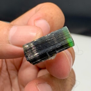 Peut inclure: Cristal de tourmaline vert foncé et noir tenu entre les doigts. Le cristal a un aspect en couches avec une teinte verte vibrante à une extrémité. Spécimen minéral naturel.