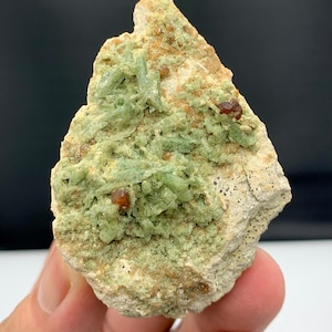 Puede incluir: Espécimen mineral natural con una formación de cristales verde claro y pequeñas inclusiones de color marrón rojizo. El mineral está en una matriz beige, con una superficie rugosa y texturizada. El espécimen se sostiene en una mano.