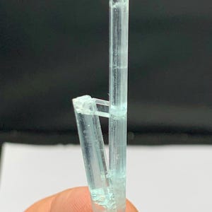 May include: A light blue aquamarine crystal cluster. The crystal is a pale, translucent blue with a vertical, elongated main crystal and a smaller, angled crystal attached. The crystal is held in a hand against a dark background.
