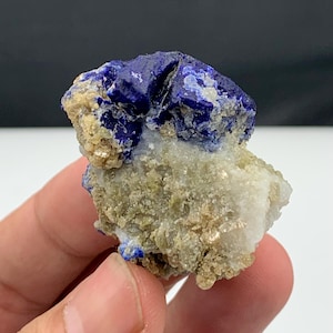 Puede incluir: Un espécimen de lapislázuli natural en bruto. El mineral azul oscuro contrasta con una matriz beige y blanca. La piedra, sostenida en la mano, muestra su textura rugosa y sin pulir, y su belleza natural.