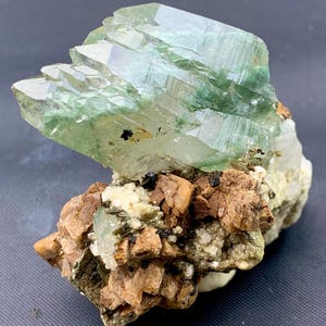 May include: A raw mineral specimen featuring a large, translucent, pale green crystal cluster perched atop a base of jagged, tan-colored rock formations with small dark inclusions and white crystalline deposits throughout the structure.